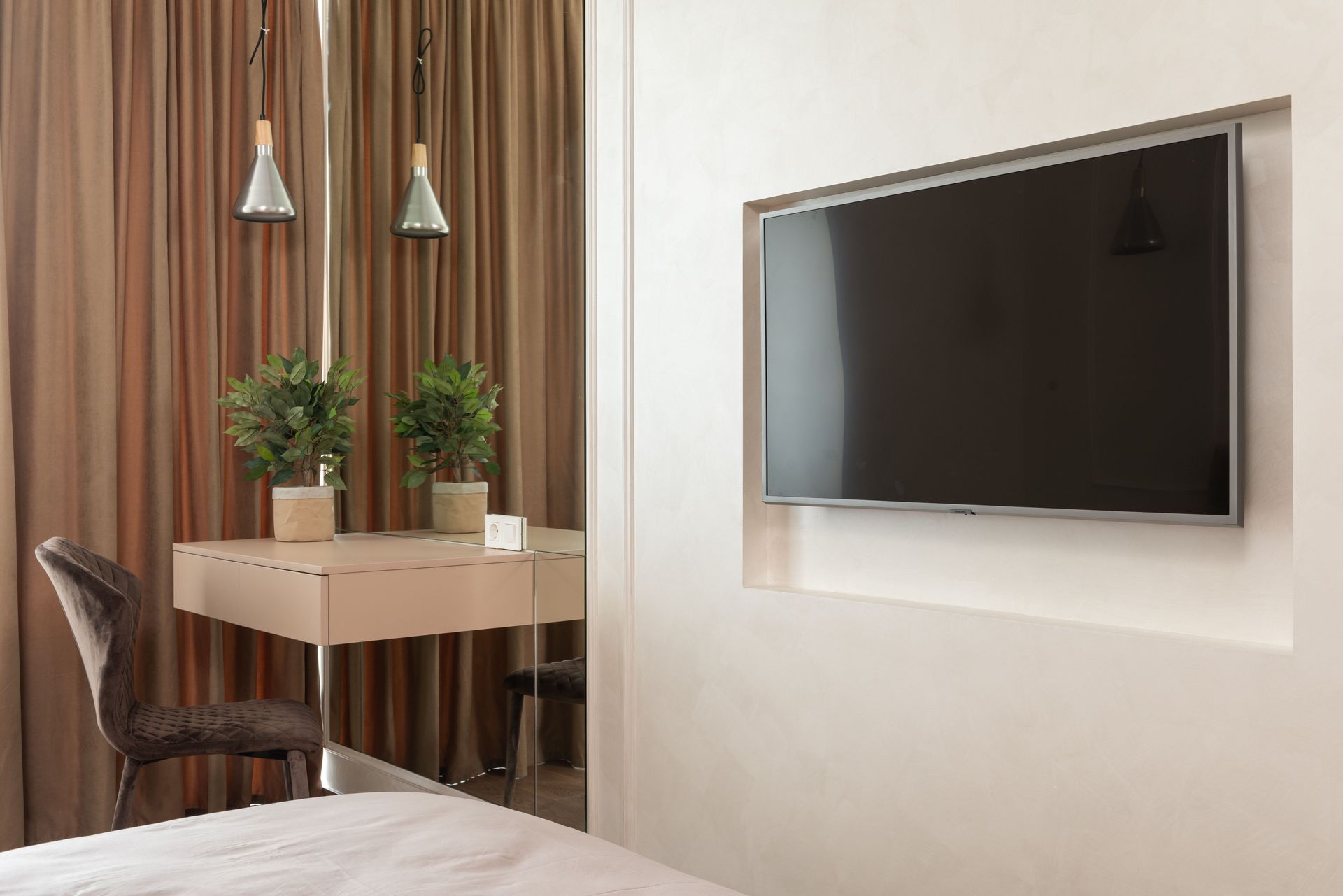 Interior of modern hotel room with TV on wall next to mirror in front of table with green potted plant and chair near curtains and bed